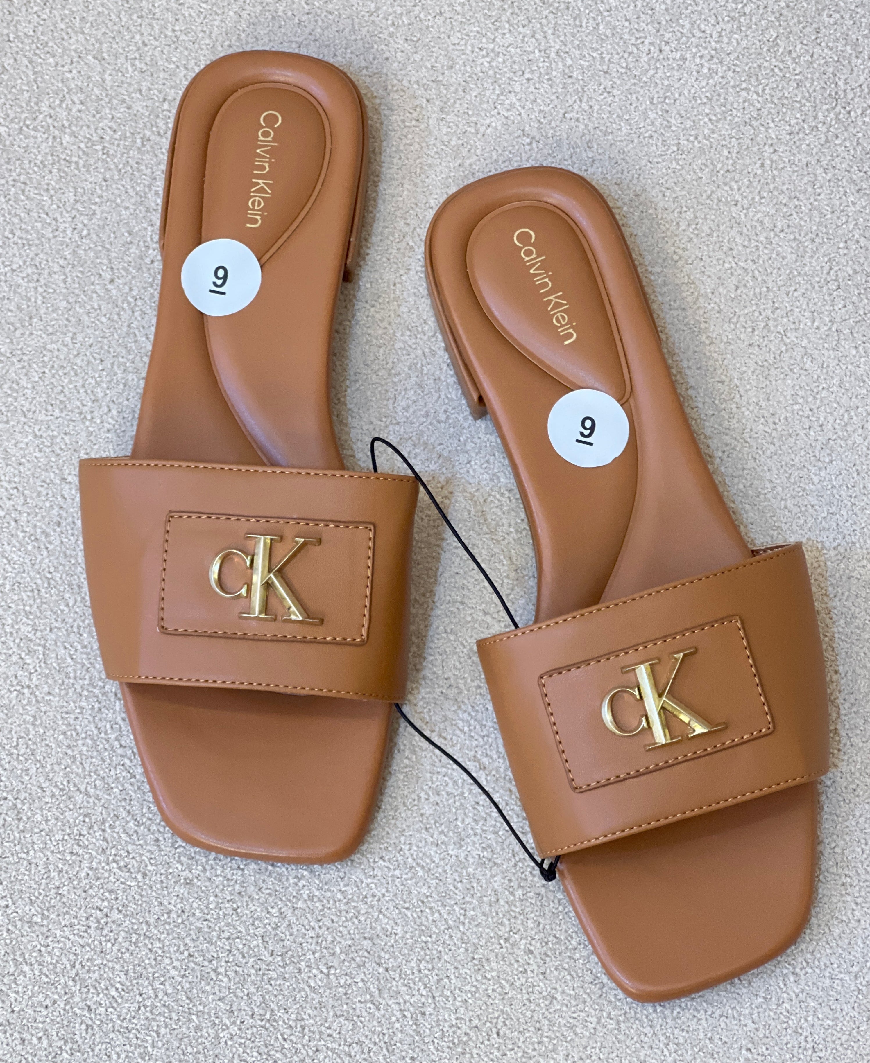 Calvin Klein slide sandals featuring a tan color, a flat heel, and a gold-toned 'ck' logo plaque on the top strap
