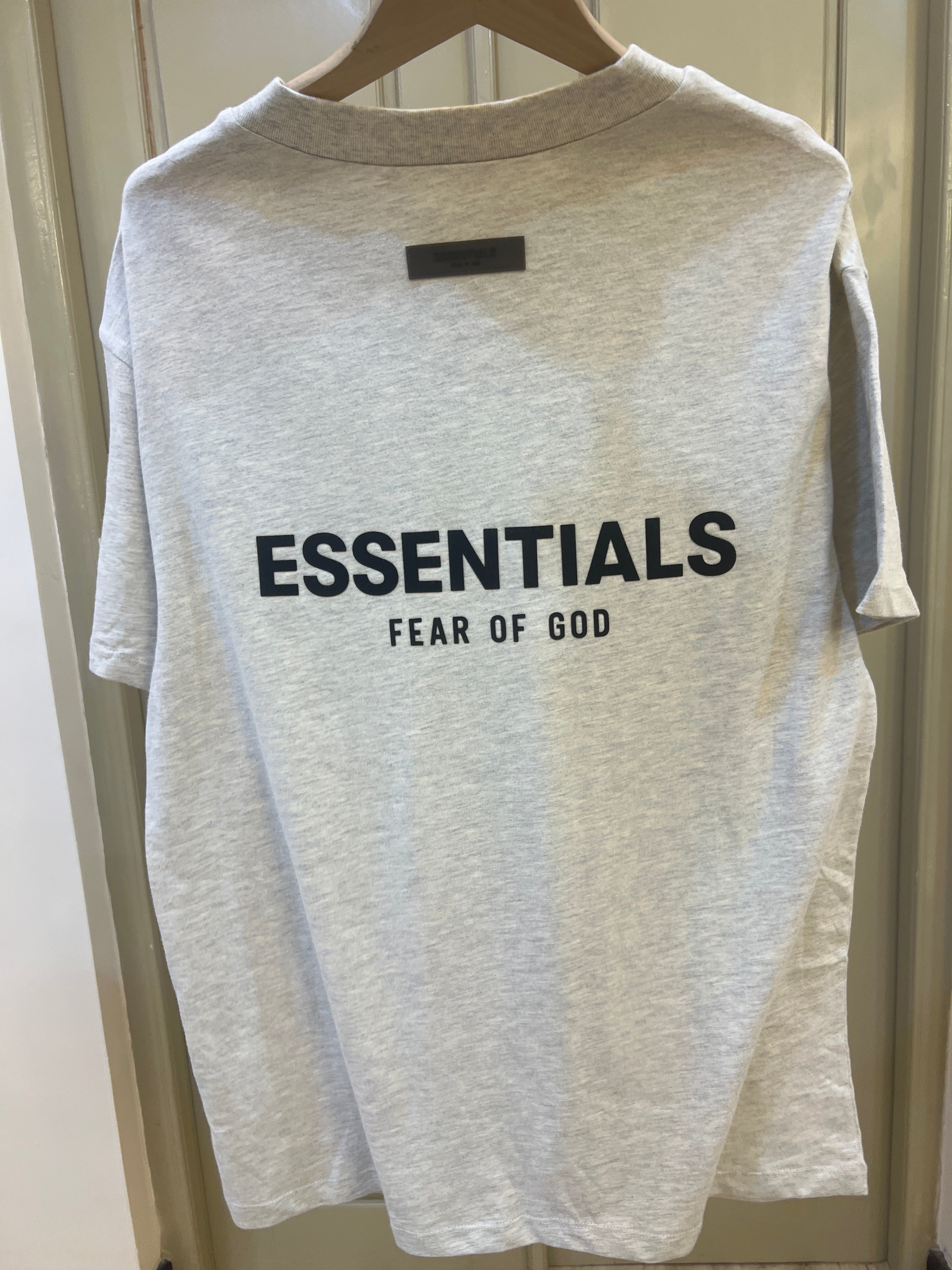 Fear of God Essentials t-shirt is rendered in a light oatmeal heather color and features a prominent branded logo on the chest