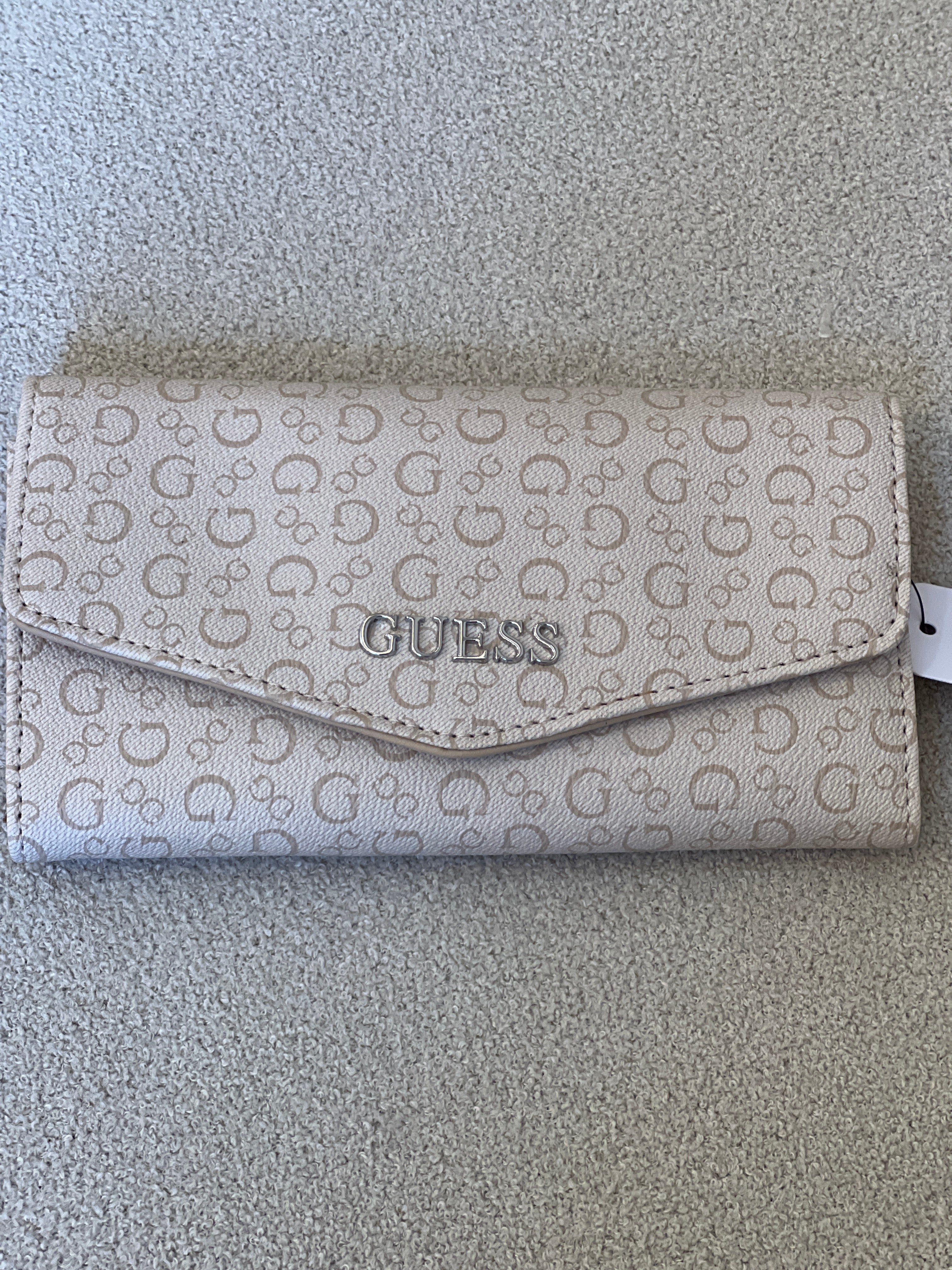Beige Guess monogram clutch wallet features a silver-tone logo emblem on the front flap