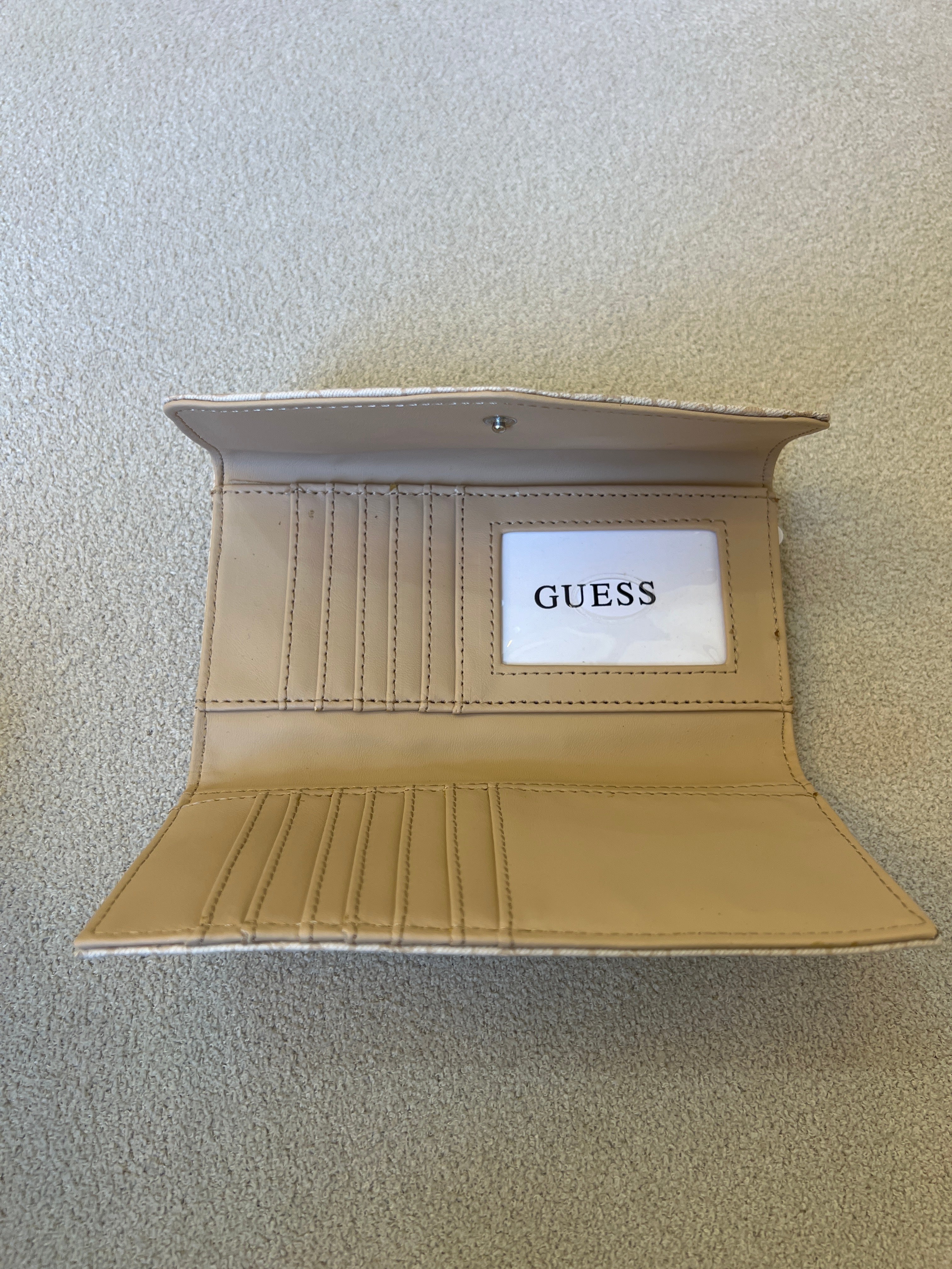 Beige Guess monogram clutch wallet features a silver-tone logo emblem on the front flap