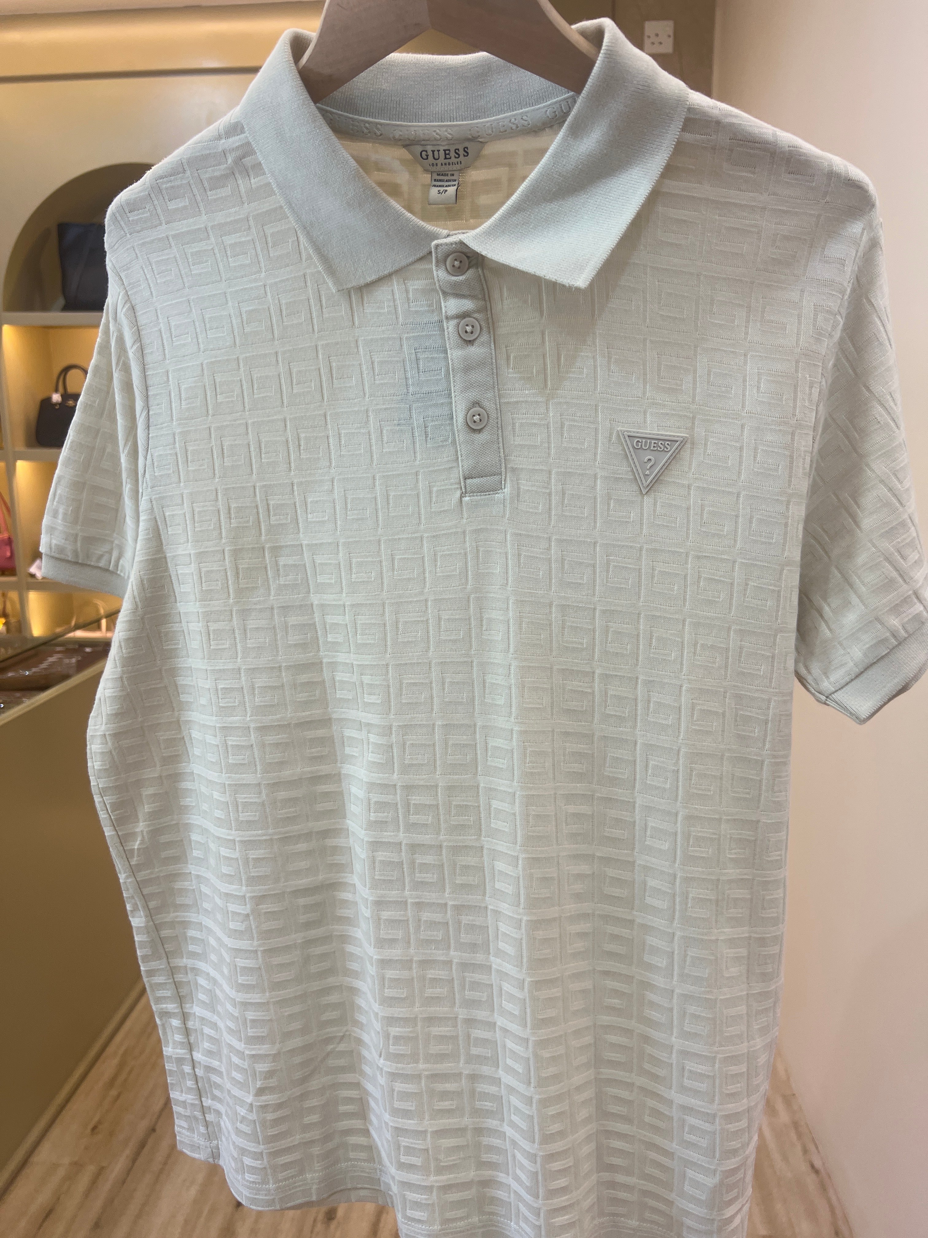 White or off-white polo shirt from the brand Guess, featuring an all-over geometric textured pattern and a small triangle logo on the chest