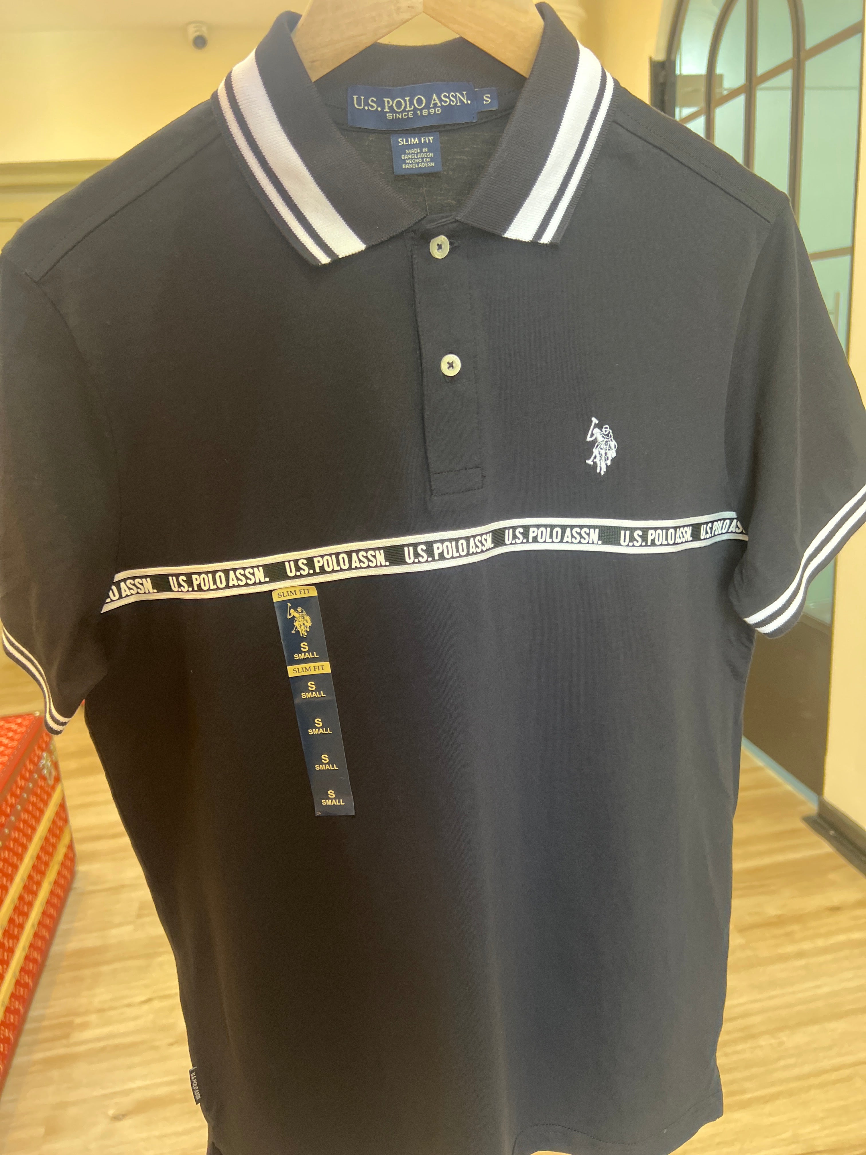 black polo shirt is a U.S. Polo Assn. men's slim fit model featuring a branded striped tape across the chest