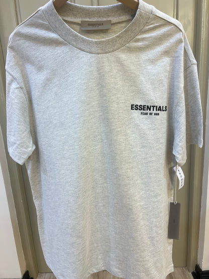 Fear of God Essentials t-shirt is rendered in a light oatmeal heather color and features a prominent branded logo on the chest